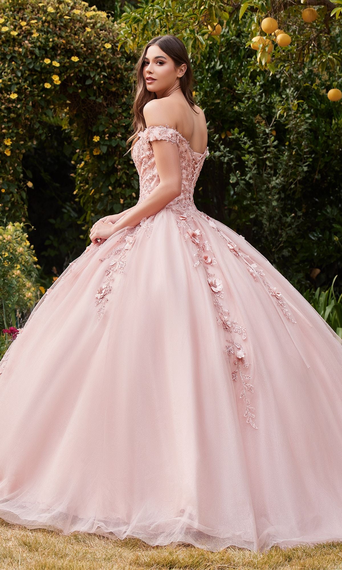 A young woman outdoors wears the Off-Shoulder Long Sweetheart Prom Ball Gown CD0185 in light pink, featuring floral-lace appliques and a full skirt. She is surrounded by lush greenery and vibrant orange trees.